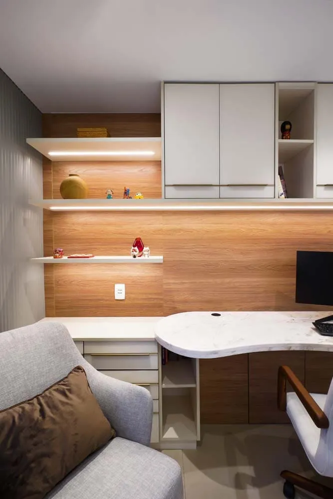 Home office com bancada de mármore branco, marcenaria amadeirada, prateleiras iluminadas, armários brancos e poltrona cinza