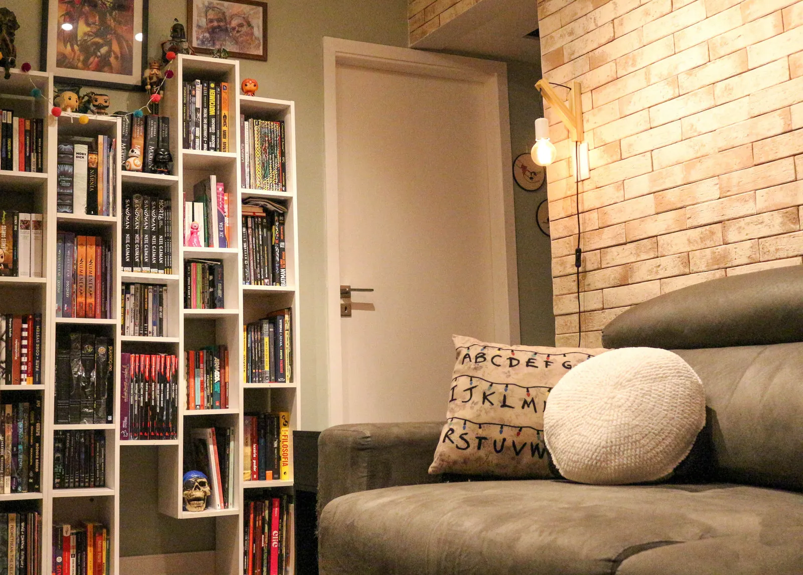 Canto de leitura com estantes brancas de livros e bonecos, sofá cinza com almofada de Stranger Things e parede de tijolinho