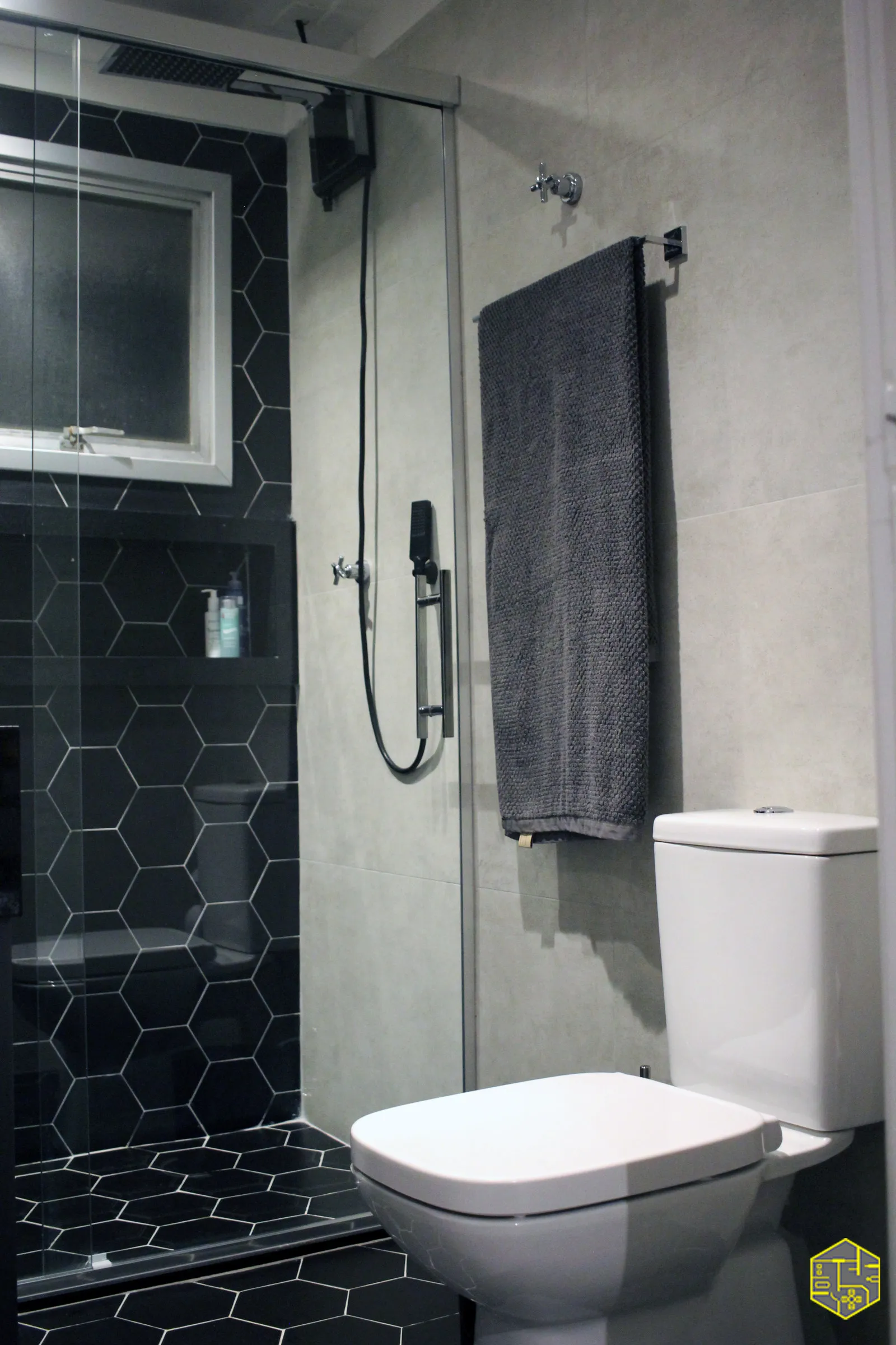 Banheiro com box de vidro, revestimento hexagonal preto no chuveiro, ducha de teto, toalha cinza e vaso sanitário branco