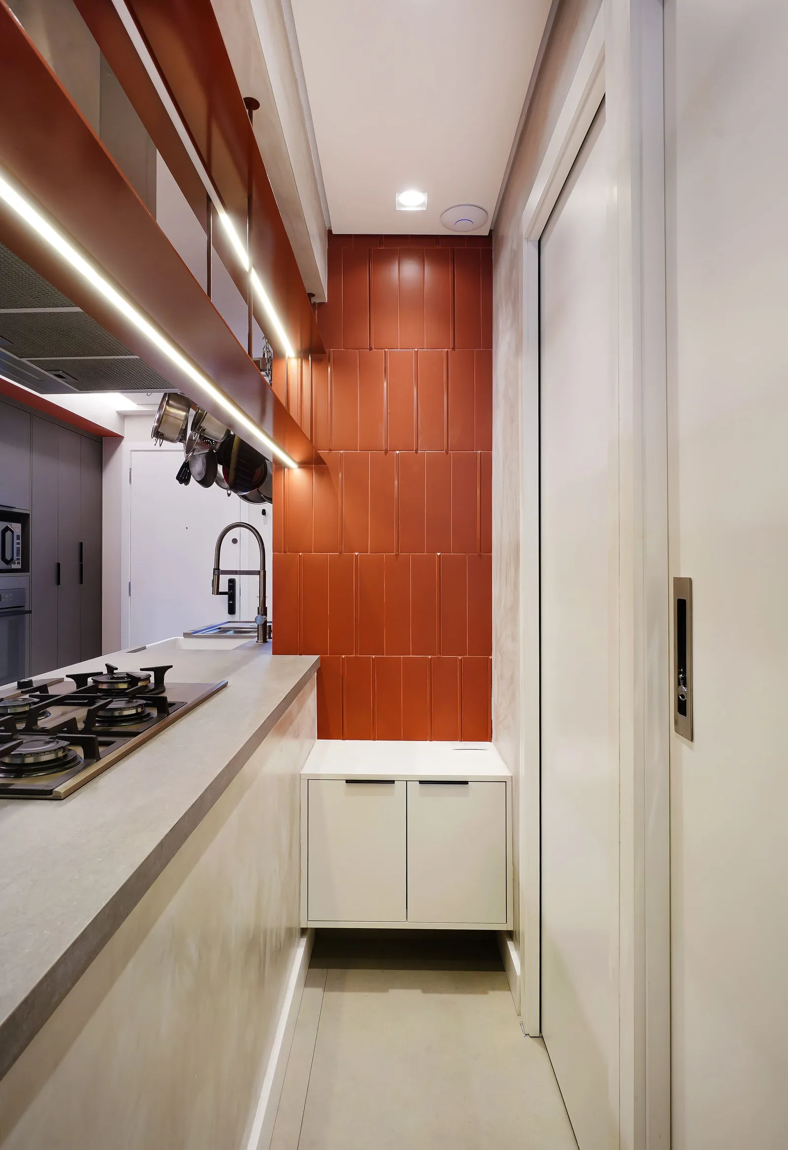 Cozinha estreita com bancada de concreto, cooktop, parede de azulejo laranja e porta de correr branca
