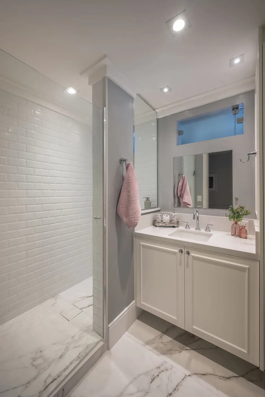 Banheiro com box de metrô branco, piso de mármore, gabinete off-white com bancada branca e toalha rosa