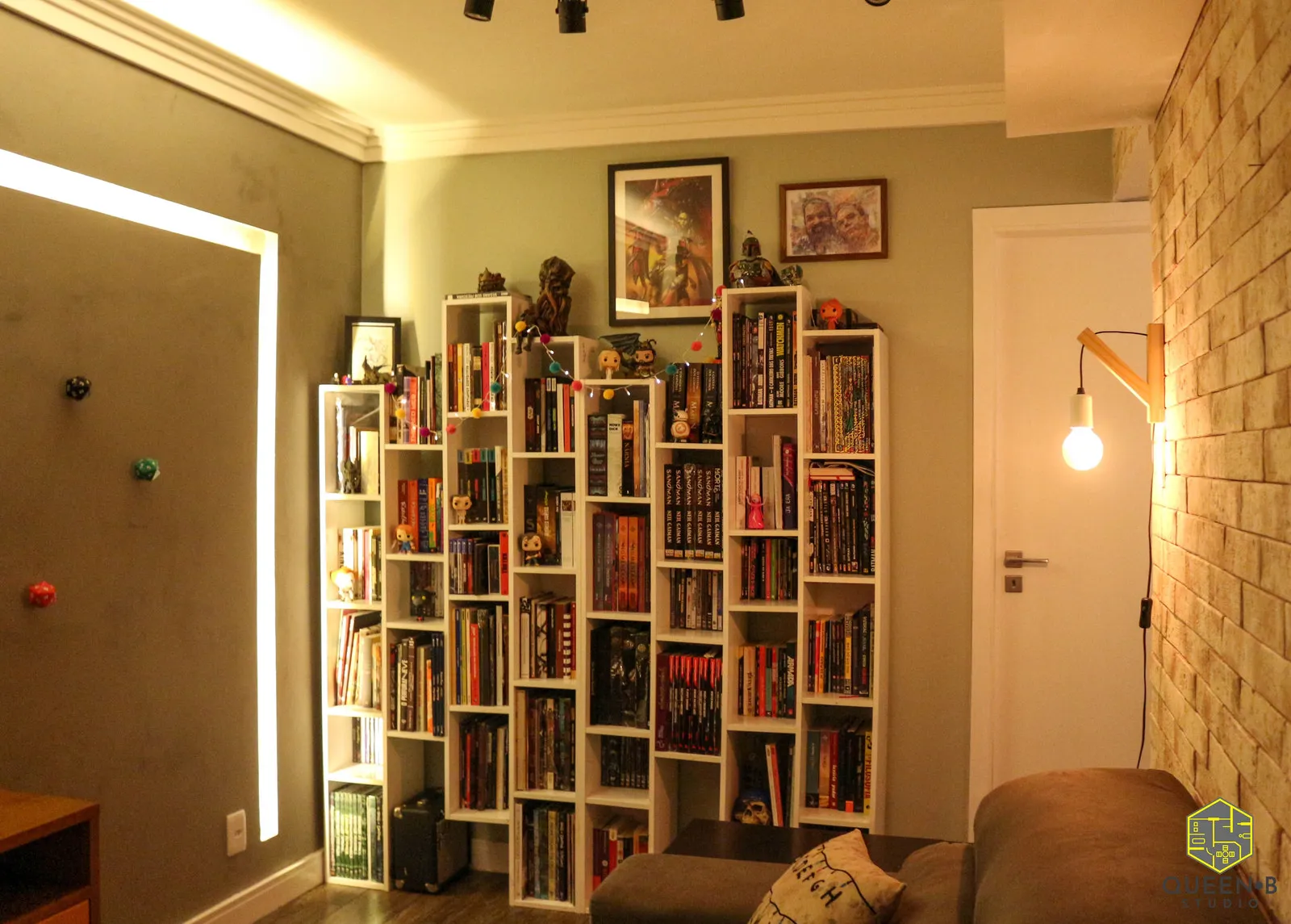 Sala com estantes brancas repletas de livros e colecionáveis, quadros emoldurados, sofá cinza e parede de tijolinho ao lado