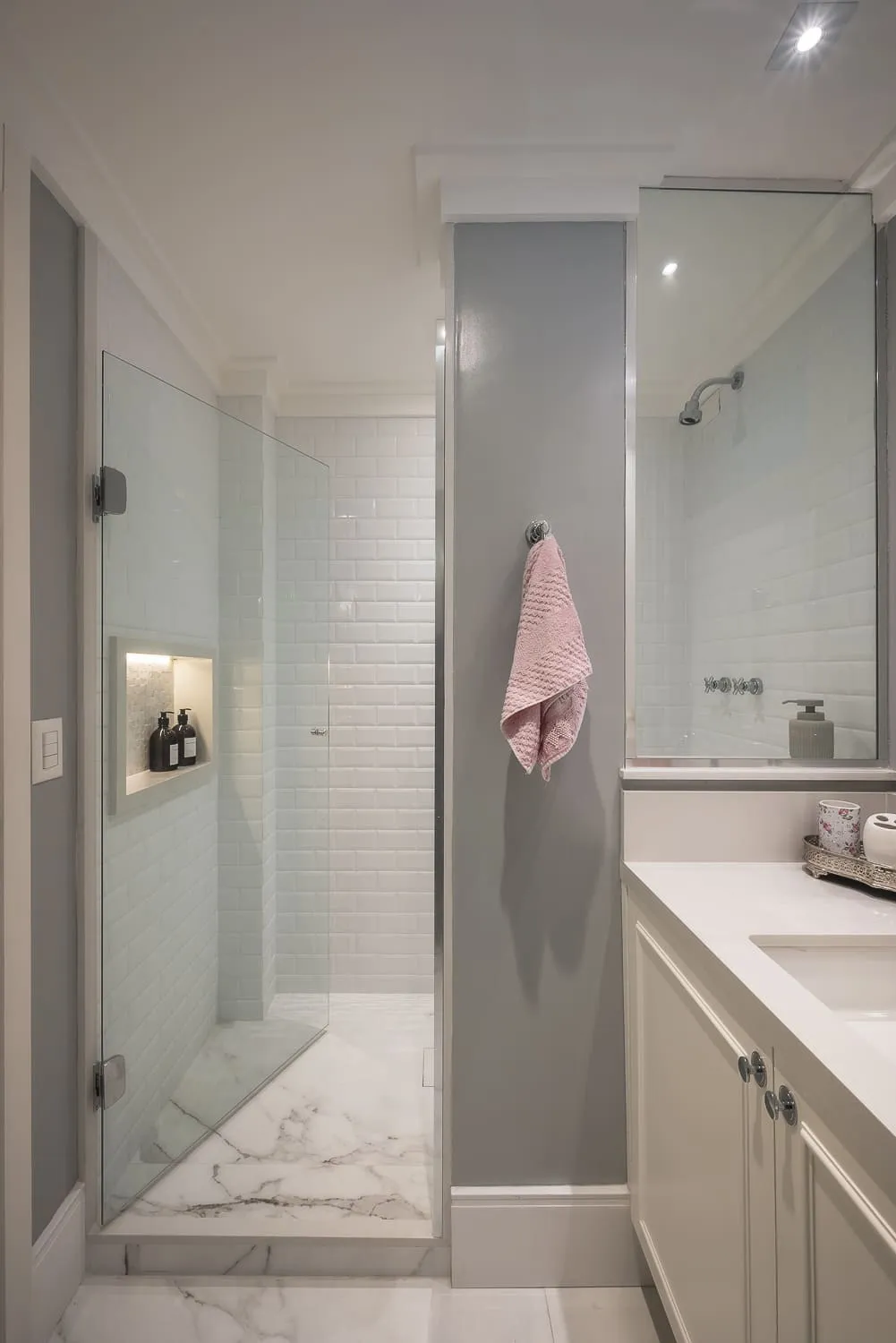 Banheiro com box de vidro, revestimento de metrô branco, nicho iluminado com produtos e piso de mármore
