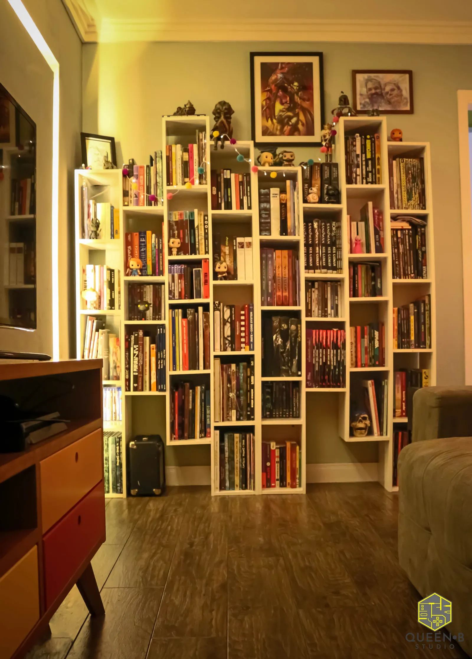 Vista ampla da sala com estantes brancas altas cheias de livros e bonecos, rack colorido retrô e piso de madeira