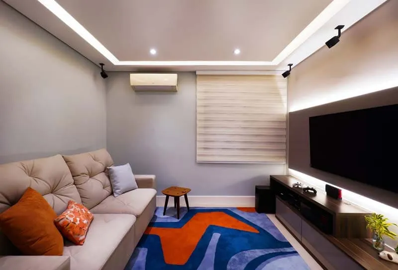 Sala de TV com sofá bege, almofadas laranja, tapete azul e laranja, painel de TV e iluminação de LED