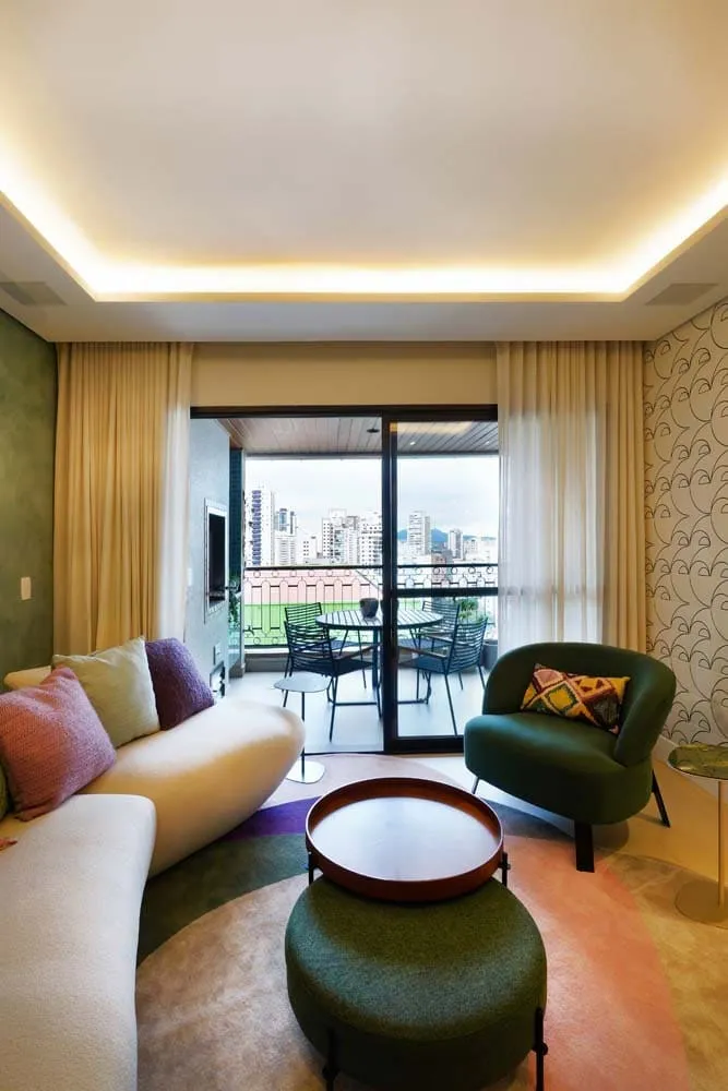 Sala de estar com sofá curvo claro, poltrona e pufe verdes, tapete colorido e varanda envidraçada com vista da cidade