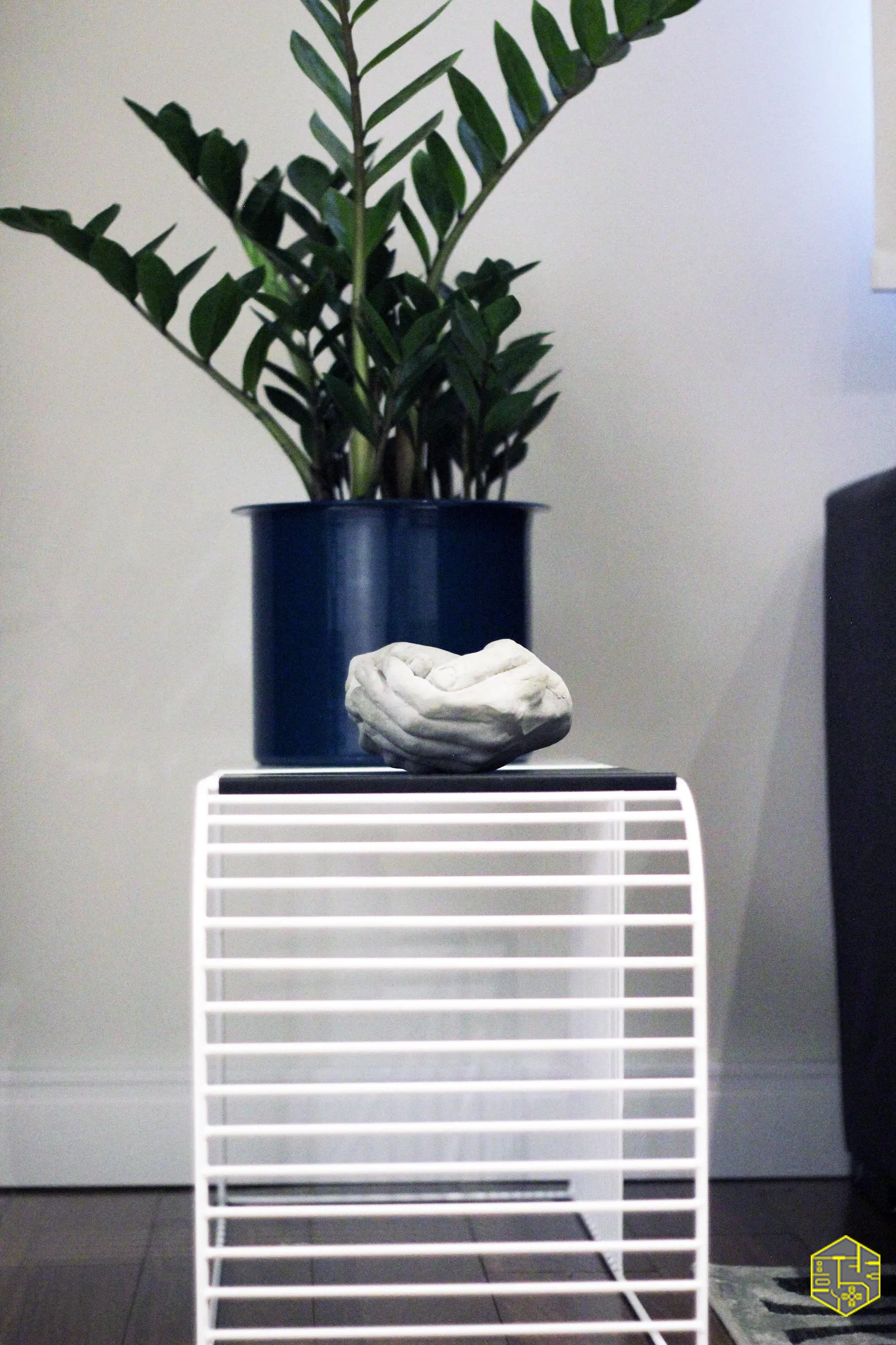 Detalhe de planta ZZ em vaso azul sobre mesa lateral de aramado branco, com escultura de pedra clara