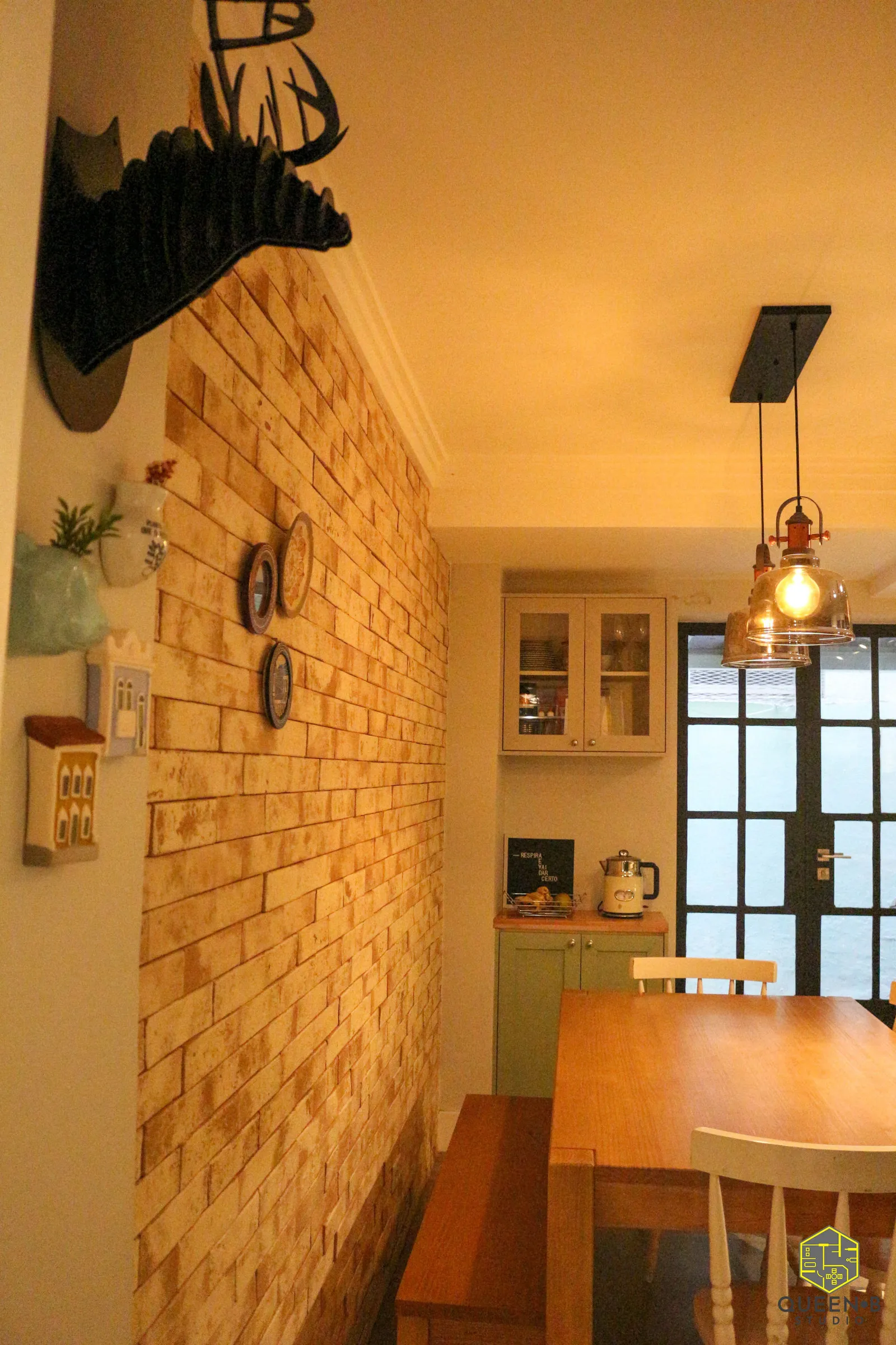 Parede de tijolinho com pratos decorativos e quadrinhos, aparador verde com chaleira e pendente de vidro na cozinha
