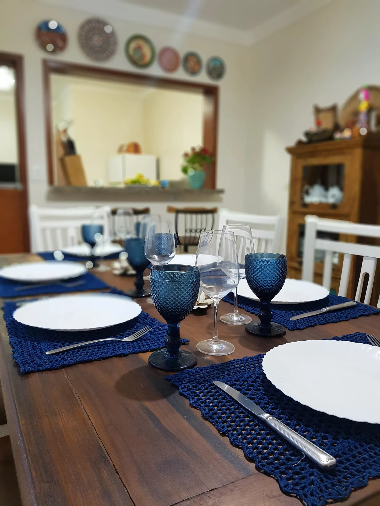 Mesa de jantar posta com taças azuis, louça branca e jogos americanos de crochê, com a passagem para a cozinha ao fundo