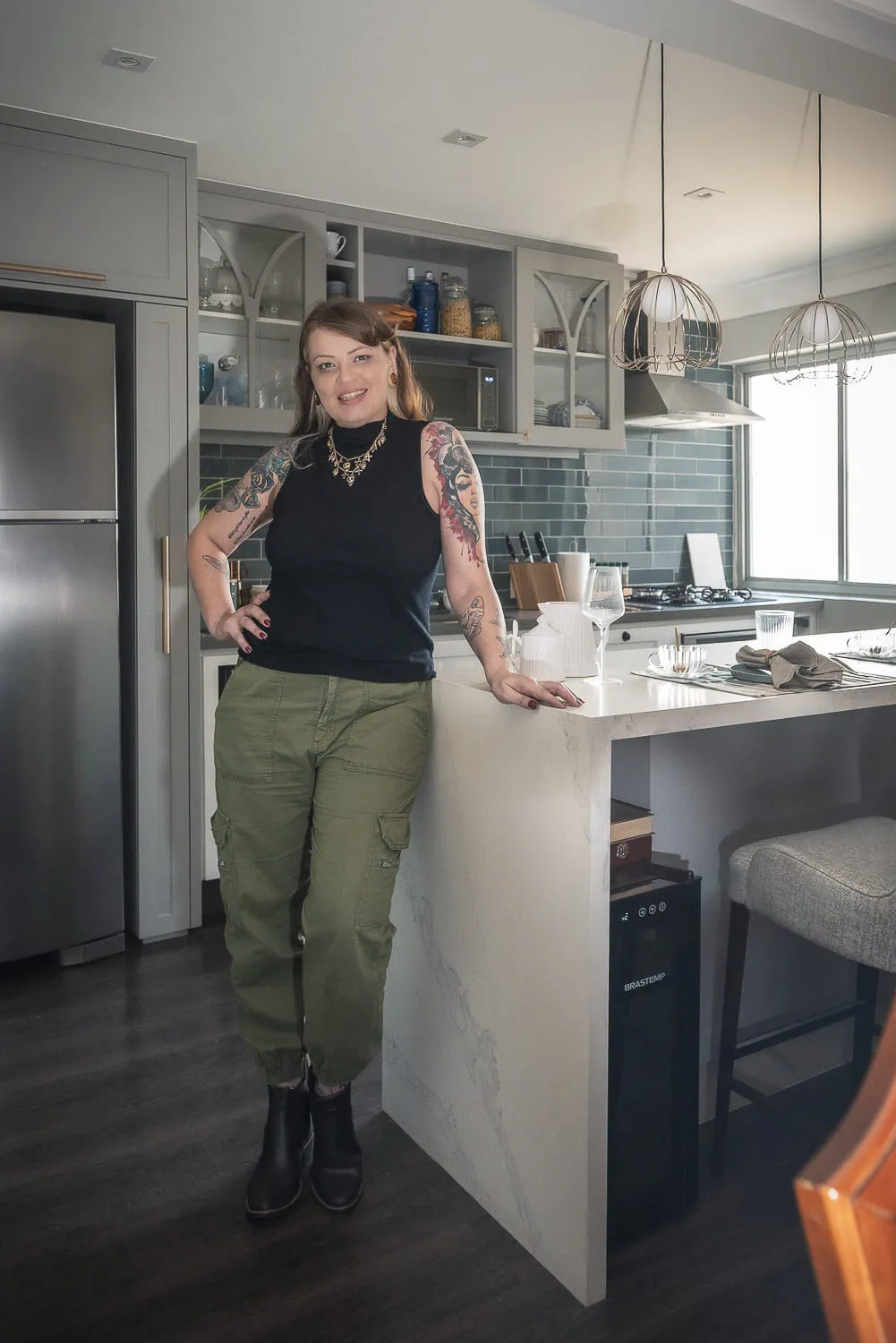 Moradora tatuada de blusa preta e calça verde apoiada na bancada de mármore da cozinha, com marcenaria cinza ao fundo