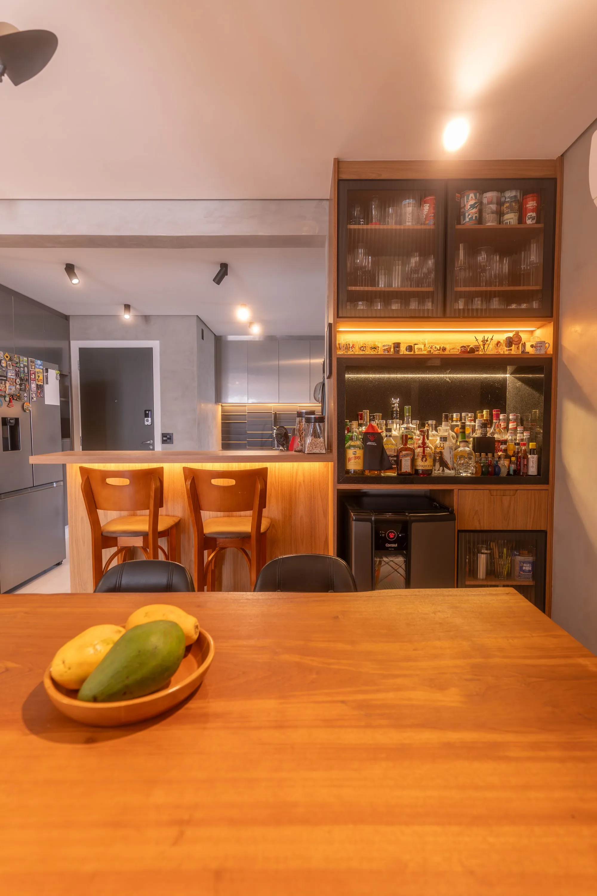 Mesa de jantar de madeira maciça com bar e adega de vidro iluminados ao lado, cozinha ao fundo