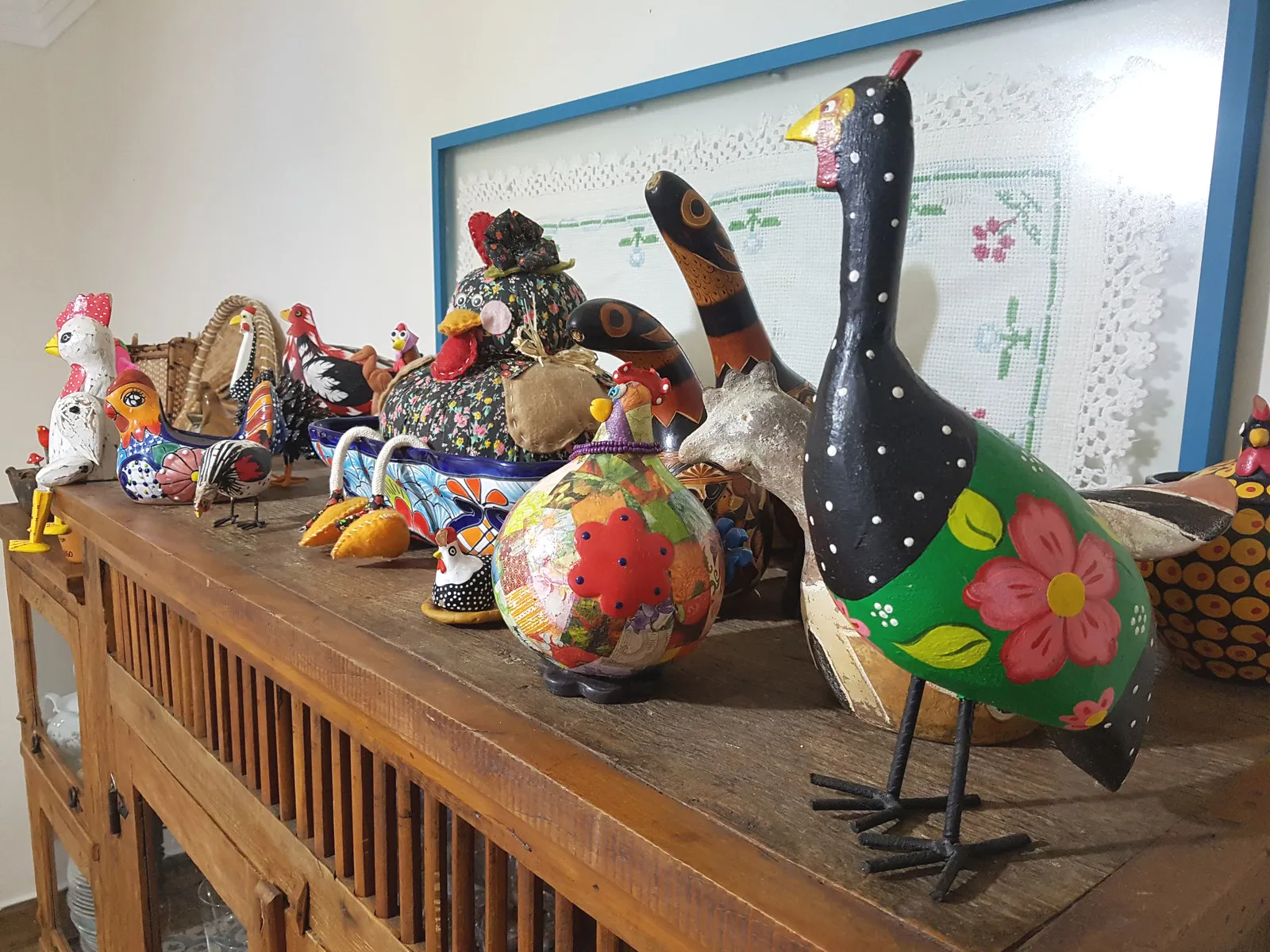 Detalhe da coleção de galos e aves de cerâmica sobre o aparador, com o quadro de bordado emoldurado de azul