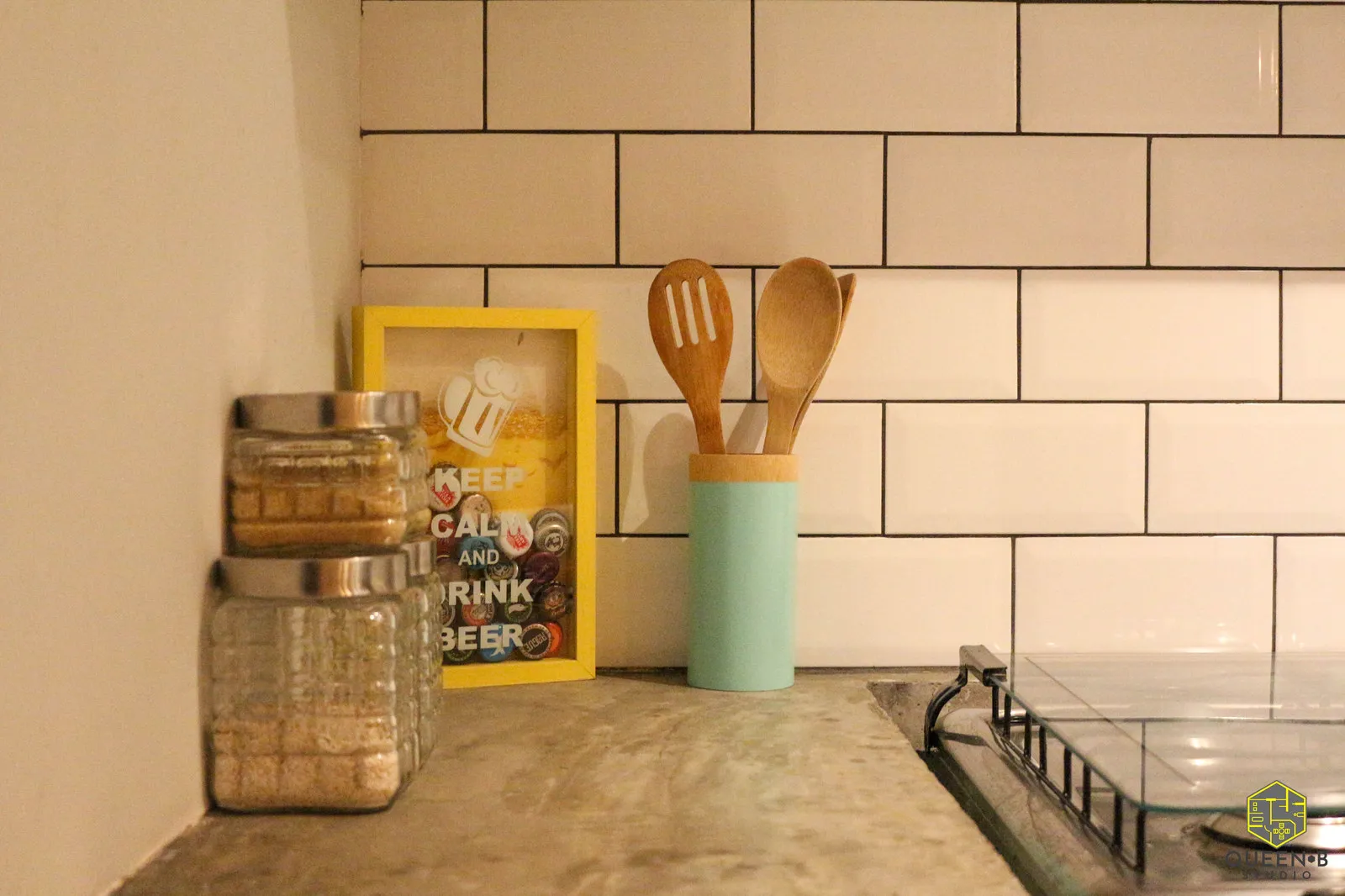 Detalhe de bancada de concreto com azulejo de metrô branco, potes de vidro, utensílios de madeira e quadrinho amarelo