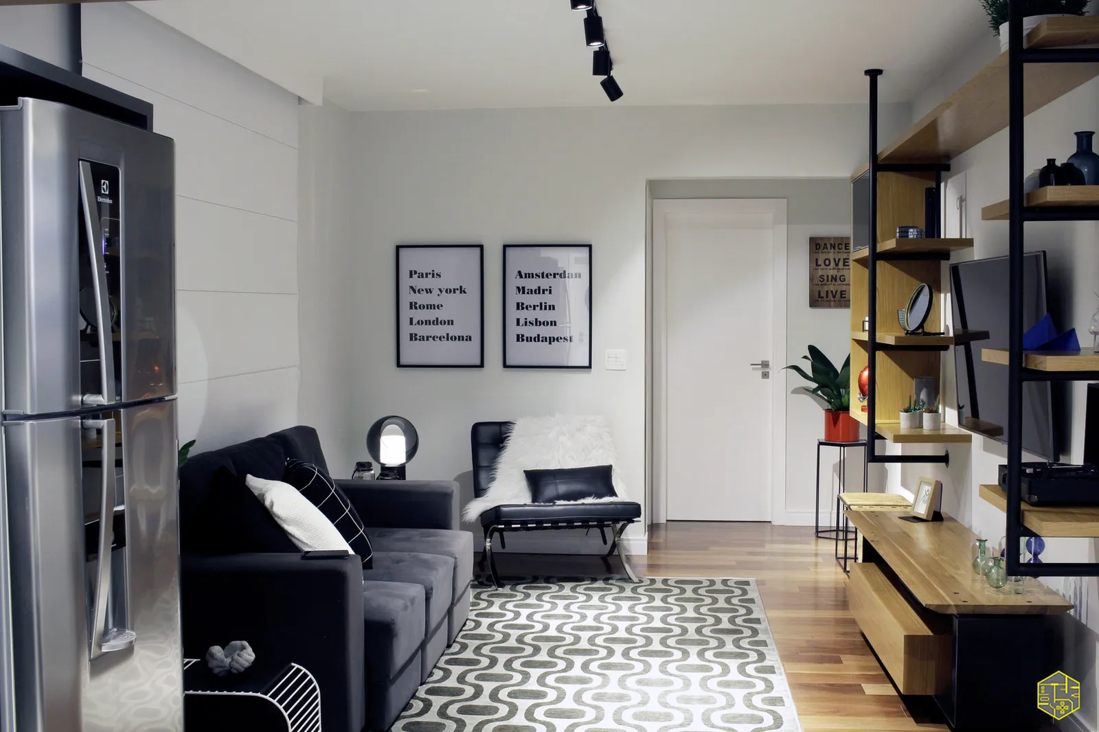 Sala integrada com geladeira inox, sofá cinza, poltrona Barcelona preta, quadros de cidades e trilho de spots pretos no teto