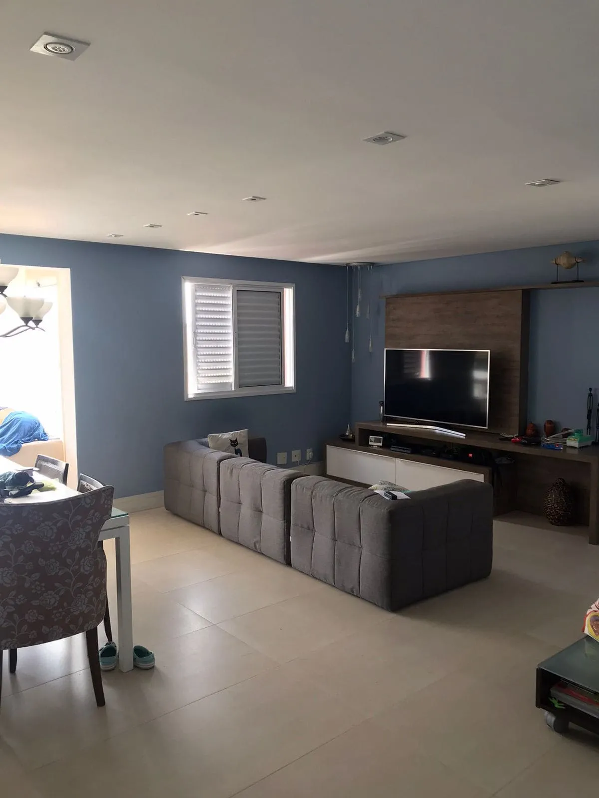 Sala de TV antes da reforma com paredes azuis, sofá cinza modular e painel de madeira, piso claro e móveis desalinhados