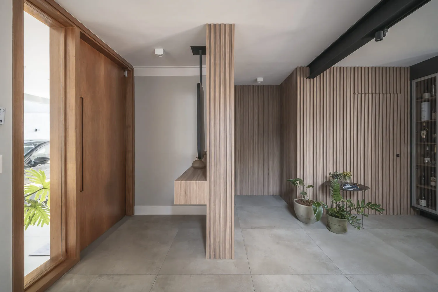 Hall de entrada com porta pivotante de madeira, parede ripada, piso cimentício e plantas junto à adega