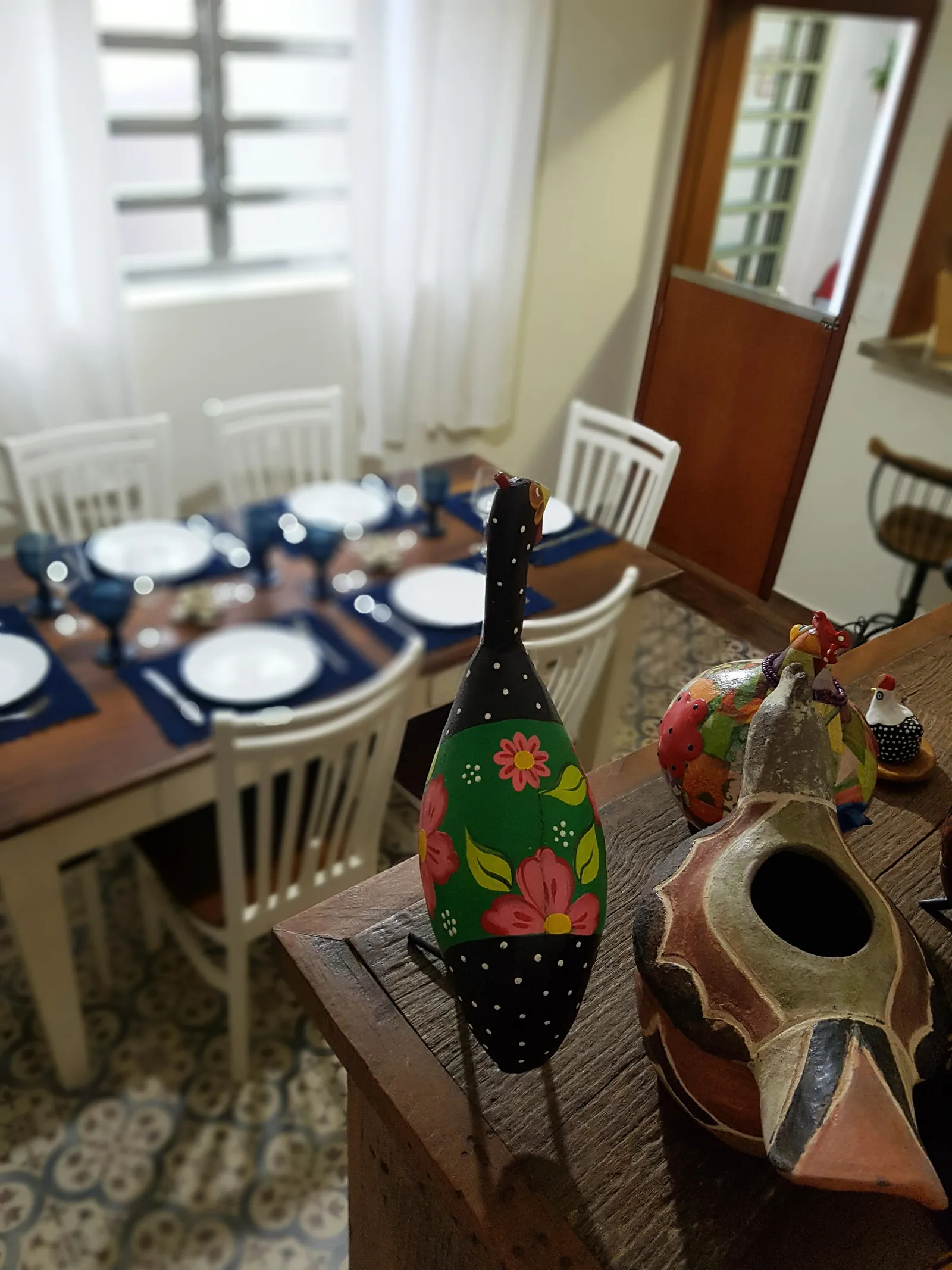 Mesa de jantar posta vista do aparador, com peças de artesanato em primeiro plano e piso de ladrilho hidráulico