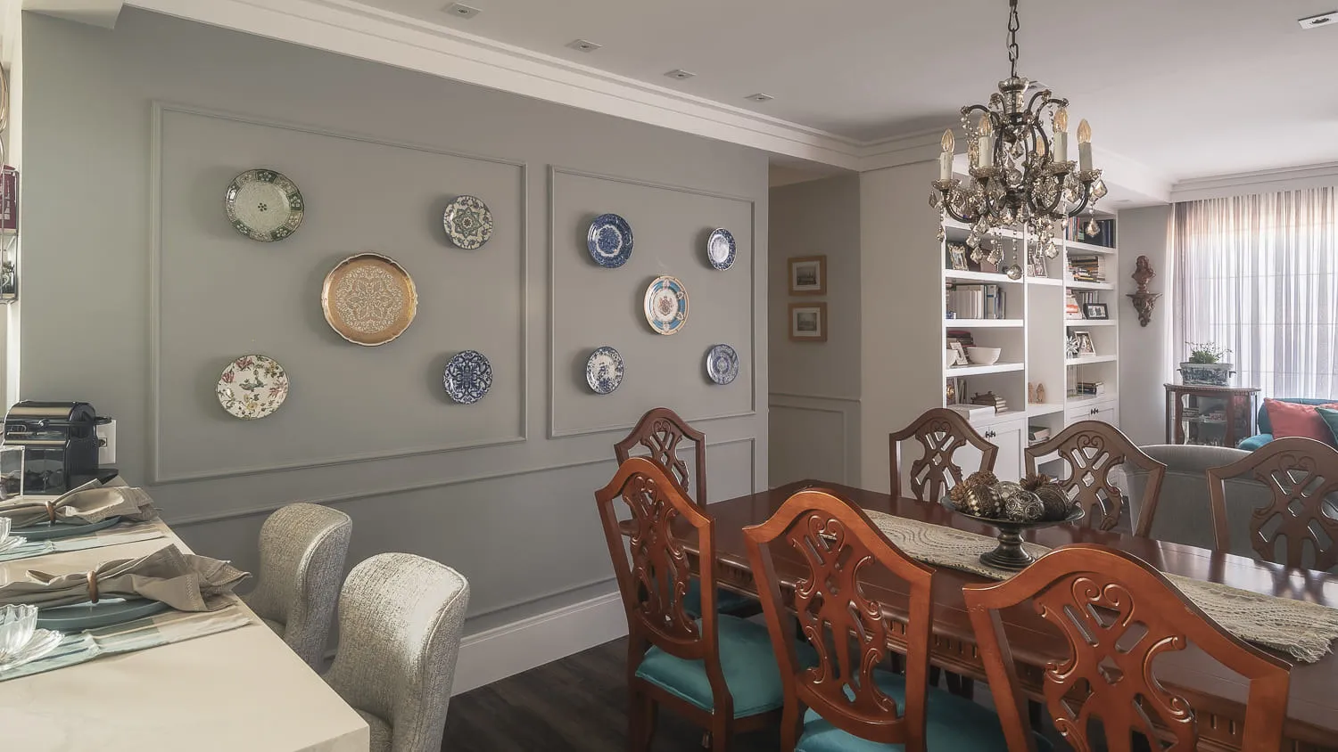Sala de jantar com mesa de madeira, cadeiras de encosto entalhado, parede cinza de pratos decorativos e lustre de cristal