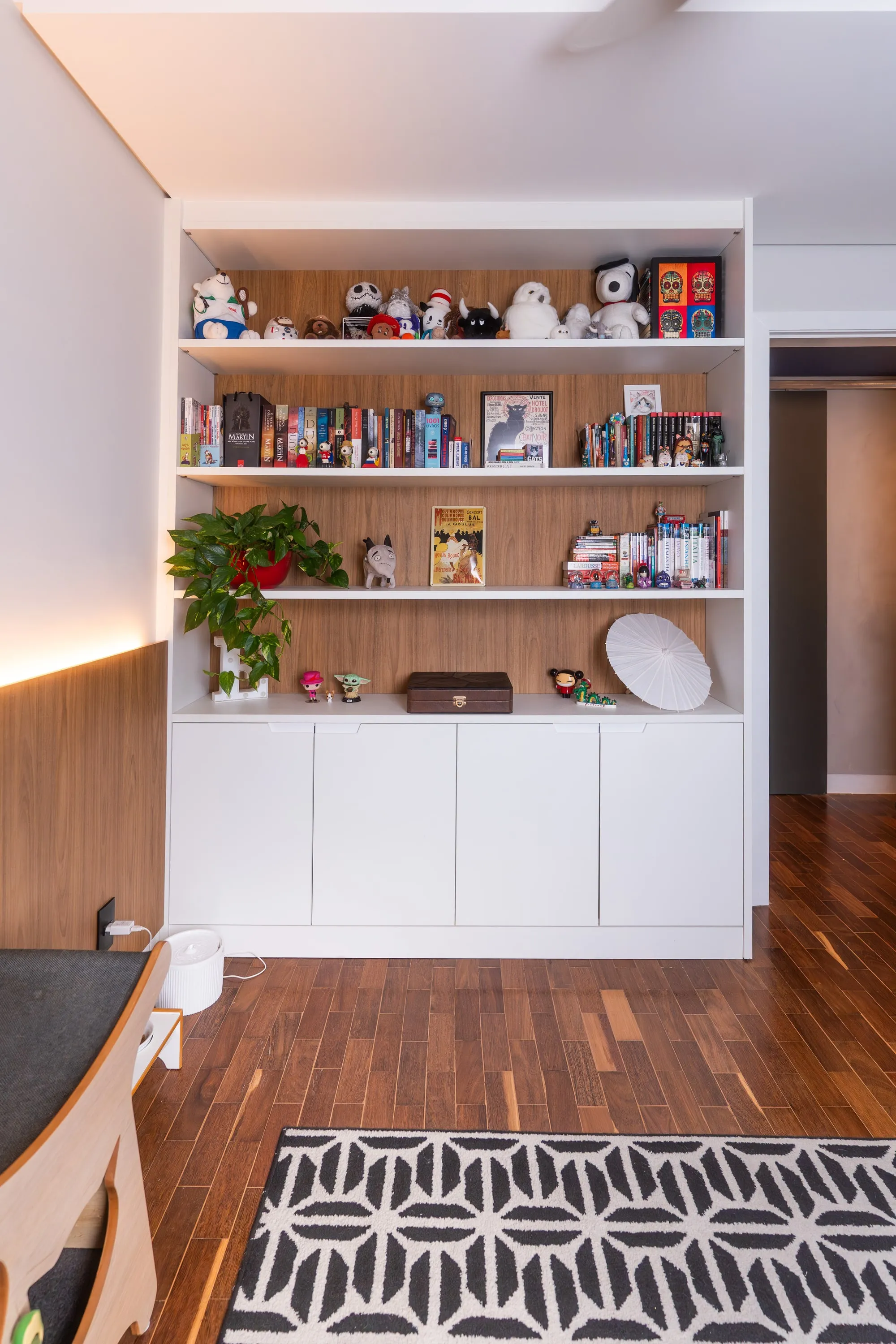 Estante branca com fundo amadeirado, pelúcias, livros e plantas, com tapete geométrico preto e branco