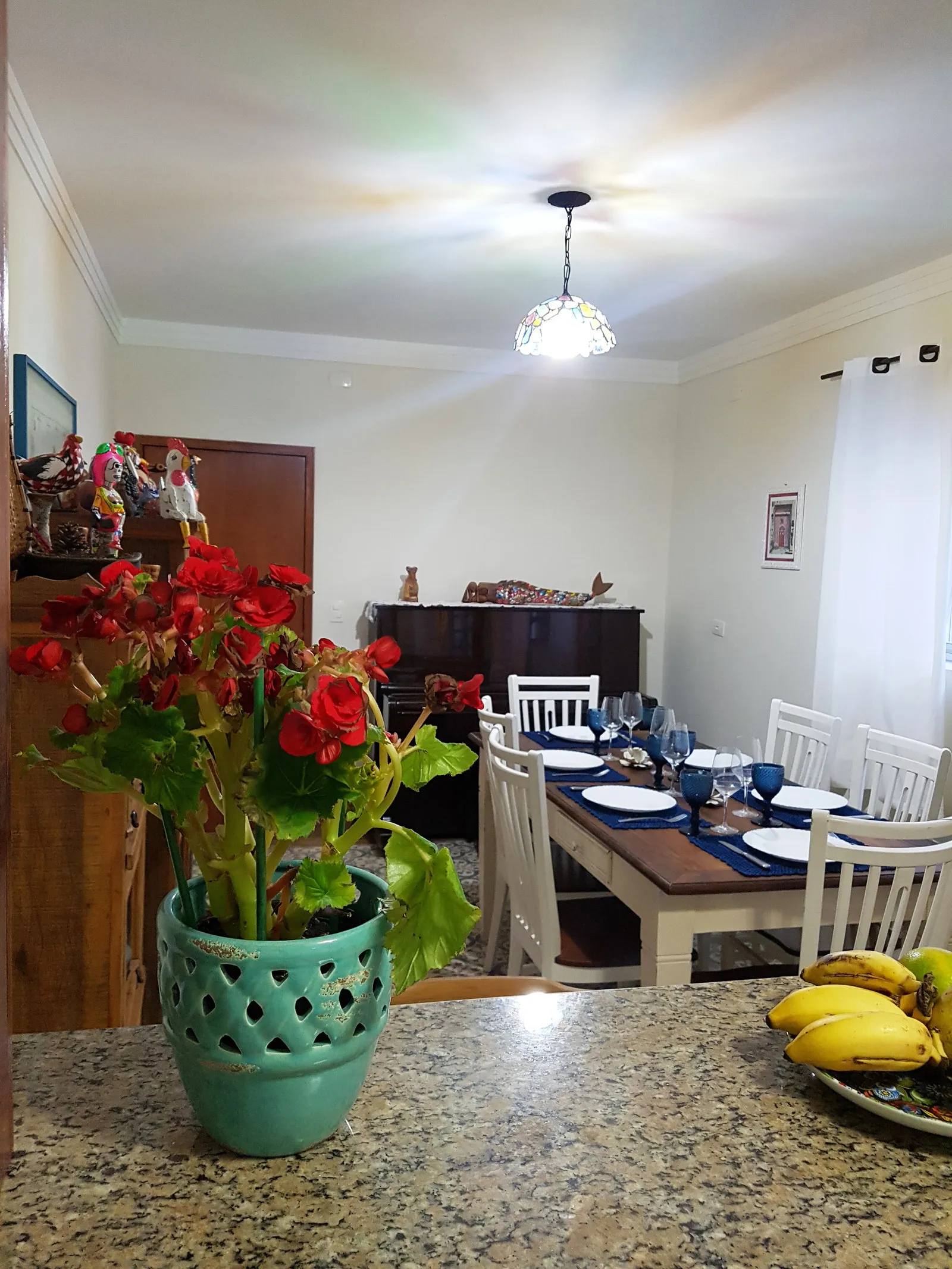 Sala de jantar com pendente de vitral, mesa posta ao fundo e vaso azul com begônias vermelhas em primeiro plano