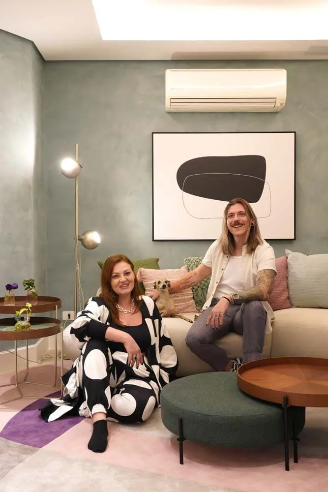 Casal e cachorro sentados no sofá da sala, com parede de cimento verde, quadro abstrato e luminária de piso