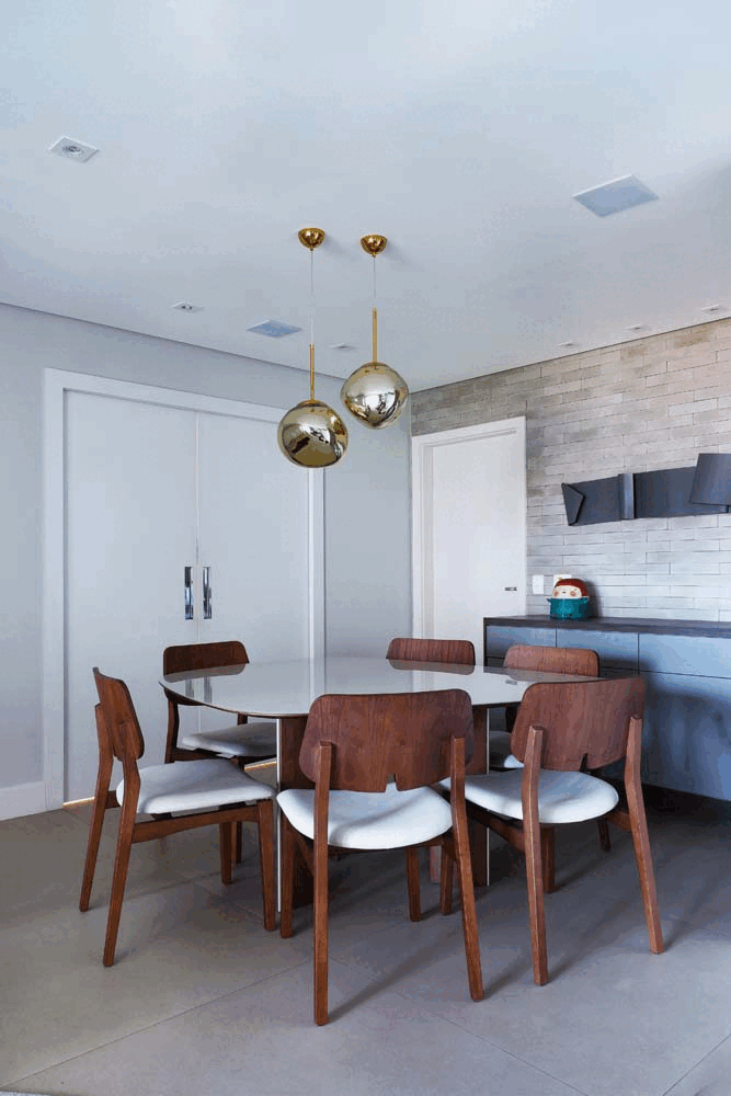 Sala de jantar com mesa oval, cadeiras de madeira, pendentes esféricos dourados e buffet cinza sobre parede de tijolinho branco