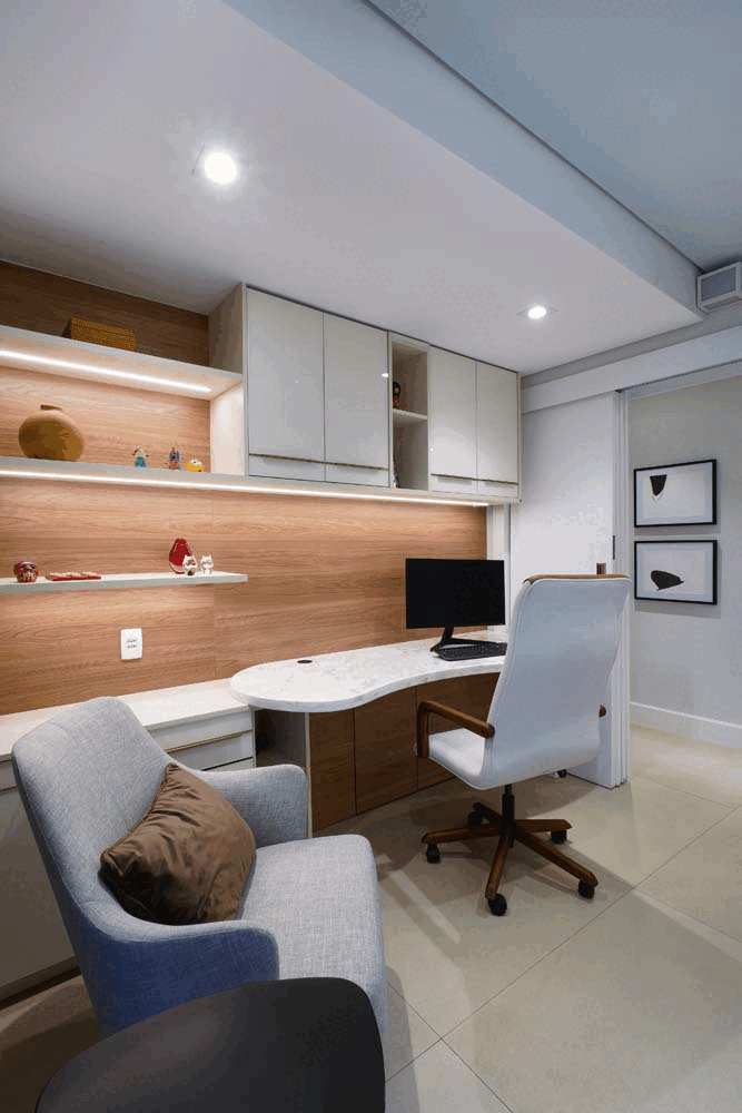 Home office com bancada de mármore orgânica, cadeira branca, poltrona cinza e prateleiras iluminadas com quadros abstratos