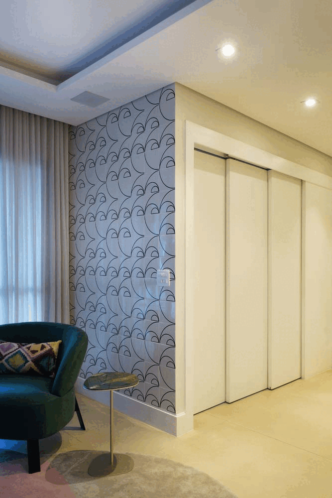 Hall de entrada com papel de parede geométrico cinza, poltrona verde e mesa lateral, ao lado de portas de correr claras