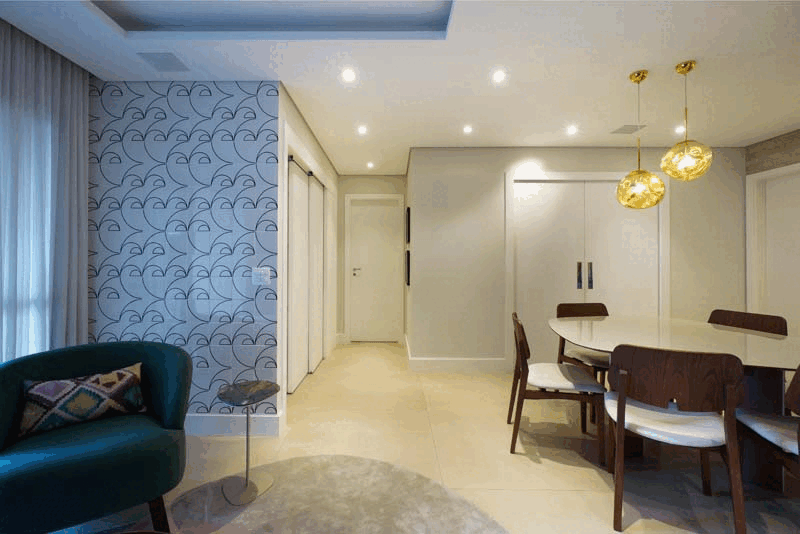 Sala de jantar integrada com mesa oval branca, cadeiras de madeira, pendentes dourados e poltrona verde junto ao papel geométrico