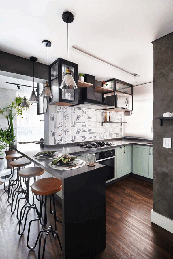 Cozinha com marcenaria verde-clara, ilha preta, revestimento geométrico preto e branco, cooktop e banquetas de madeira sobre piso amadeirado