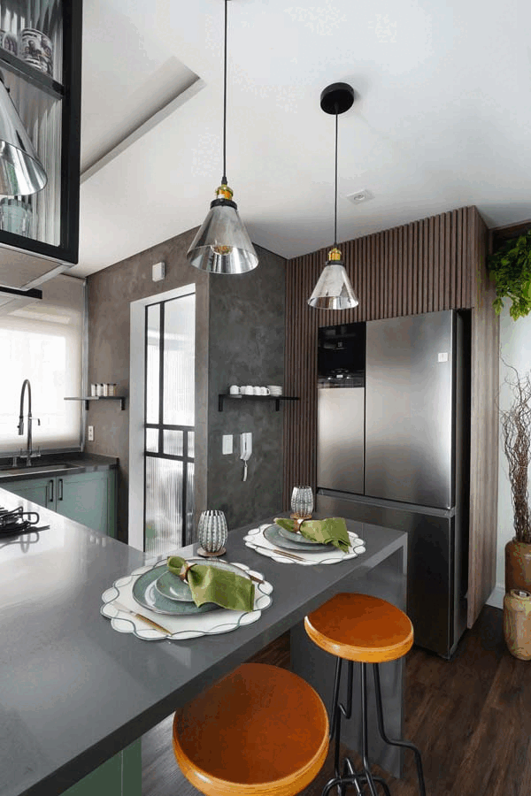 Península cinza posta com pratos e banquetas laranja, painel ripado de madeira com geladeira inox e parede de cimento queimado, sob pendentes de vidro