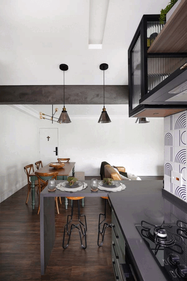 Vista da cozinha para a sala, com cooktop na bancada, mesa cinza posta e pendentes de vidro, mesa de jantar e sofá ao fundo