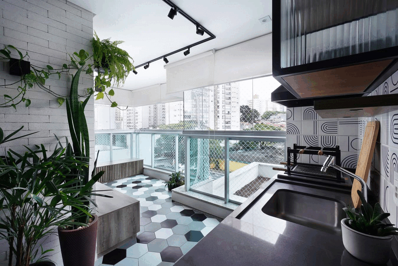 Varanda gourmet com bancada cinza e cuba, parede de tijolo branco com plantas, piso hexagonal e guarda-corpo de vidro com vista da cidade