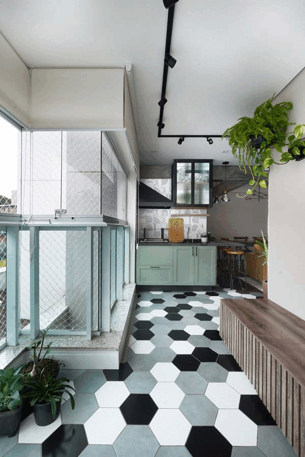 Varanda com piso hexagonal preto e branco, guarda-corpo de vidro, bancada verde-clara ao fundo e banco de madeira ripada com plantas